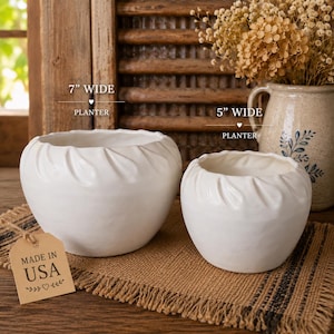 May include: Two white ceramic planters, 7 inches and 5 inches wide, with a textured leaf design around the rim. The planters are on a burlap surface, with a "Made in USA" tag. The background includes a wooden shutter and a vase of dried flowers.