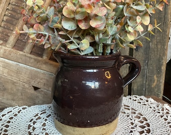 May include: A dark brown ceramic pitcher with a tan base holds a bundle of faux eucalyptus. The eucalyptus has green, red, and yellow leaves. The pitcher sits on a white crocheted doily.
