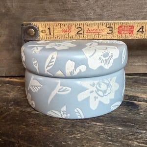 Może przedstawiać: Jasnoniebieskie, okrągłe ceramiczne pudełko z pokrywką, z białym kwiatowym wzorem. Pudełko ma średnicę około 5 cm. Pudełko stoi na drewnianej powierzchni.