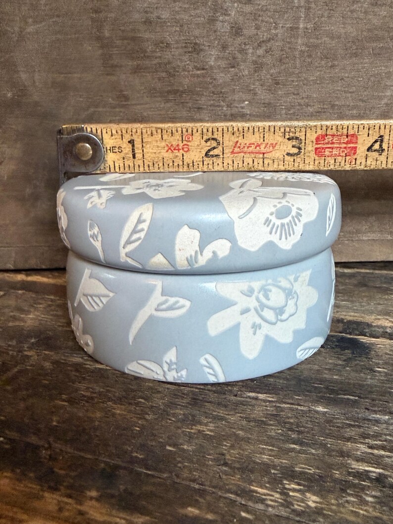 Może przedstawiać: Jasnoniebieskie, okrągłe ceramiczne pudełko z pokrywką, z białym kwiatowym wzorem. Pudełko ma średnicę około 5 cm. Pudełko stoi na drewnianej powierzchni.