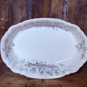 May include: An oval ceramic serving platter with a white base and a scalloped edge. The platter features a detailed grey botanical transfer print of thistle flowers and foliage along the rim and the bottom interior surface.