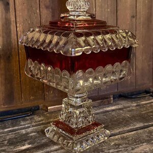 May include: A square glass candy dish with a deep red center band and clear, textured glass edges. The piece features a matching square lid with a decorative finial top, all supported by a sturdy, square pedestal base with intricate detailing.