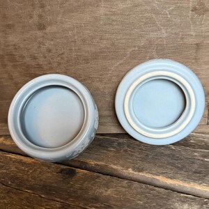 Może przedstawiać: Jasnoniebieski ceramiczny słoik z pasującą pokrywką. Słoik jest okrągły z kwiatowym wzorem z boku. Pokrywka jest r&oacute;wnież okrągła z zagłębionym środkiem. Słoik i pokrywka leżą na drewnianej powierzchni.