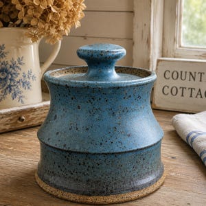 May include: A blue ceramic canister with a lid, featuring a speckled finish and a flared silhouette. The base is unglazed, showing a natural tan texture. The body transitions from a lighter blue at the top to a darker, deeper blue at the bottom.