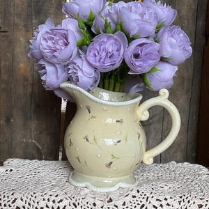 May include: A cream-colored pitcher with a floral design holds a bouquet of lavender roses. The pitcher has a scalloped rim and handle. The arrangement sits on a white, crocheted doily, with a rustic wooden background.