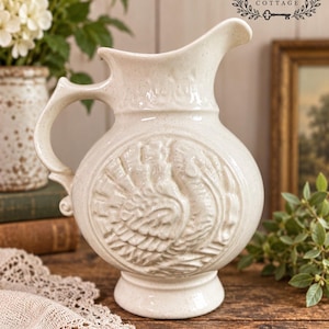 May include: A cream-colored ceramic pitcher featuring a speckled finish and a prominent embossed relief of a turkey on the front. The vessel has a curved handle, a wide spout, and a circular base, with the text THE FOX KEY COTTAGE visible.