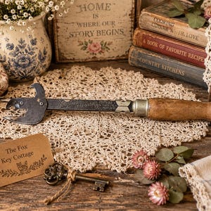 May include: A vintage metal can opener with a wooden handle and a dark finish. The metal shaft features engraved text including the word Vanguard. The tool is positioned horizontally, showcasing its functional cutting head and a smooth, rounded grip.