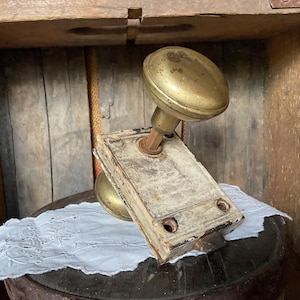 May include: An antique brass doorknob set with a rectangular backplate. The doorknob is a classic oval shape, showing signs of age and wear. The backplate has a distressed, off-white finish. The set is likely from a vintage home.
