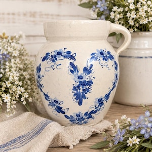 May include: A cream-colored stoneware pitcher with a blue floral design. The pitcher has a handle and is decorated with blue flowers and leaves. A linen towel with blue stripes and flowers are in the background.