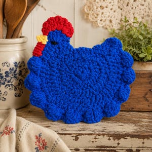 May include: A crochet potholder shaped like a chicken, featuring a vibrant blue body, a textured red comb, and a small yellow beak. The piece is crafted with a dense, looped stitch pattern that creates a rounded, scalloped edge along the perimeter.