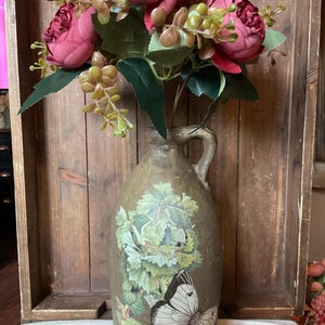 May include: A rustic, olive-green ceramic jug with a handle, adorned with a butterfly and leaf design. The jug holds a bouquet of faux burgundy peonies and greenery. The overall aesthetic is vintage and farmhouse-inspired.