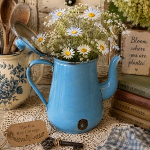 May include: A bright blue enamel teapot with a hinged lid, filled with white daisies and baby's breath. A brown paper tag reads The Fox Key Cottage, accompanied by a metal skeleton key. The teapot features a curved handle and spout.