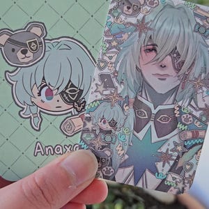 Two of the same photocard are shown. One to display the back-faced card, while the other displays the front face of the card. It is of the character Anaxa from honkai star rail. The back-face contains a chibi version of the character while the front is a more detailed illustration of said character decorated with stickers all around them.