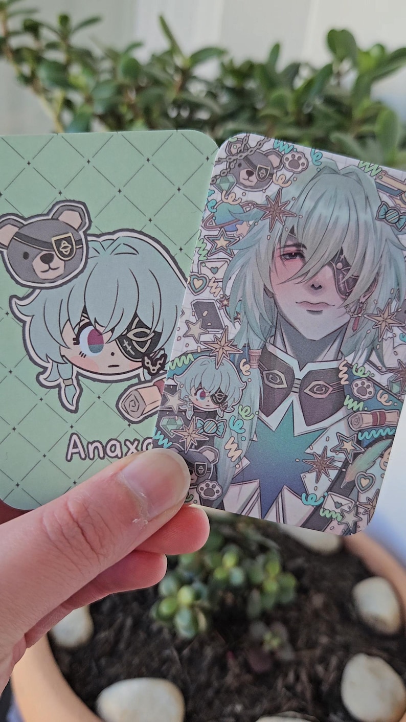 Two of the same photocard are shown. One to display the back-faced card, while the other displays the front face of the card. It is of the character Anaxa from honkai star rail. The back-face contains a chibi version of the character while the front is a more detailed illustration of said character decorated with stickers all around them.