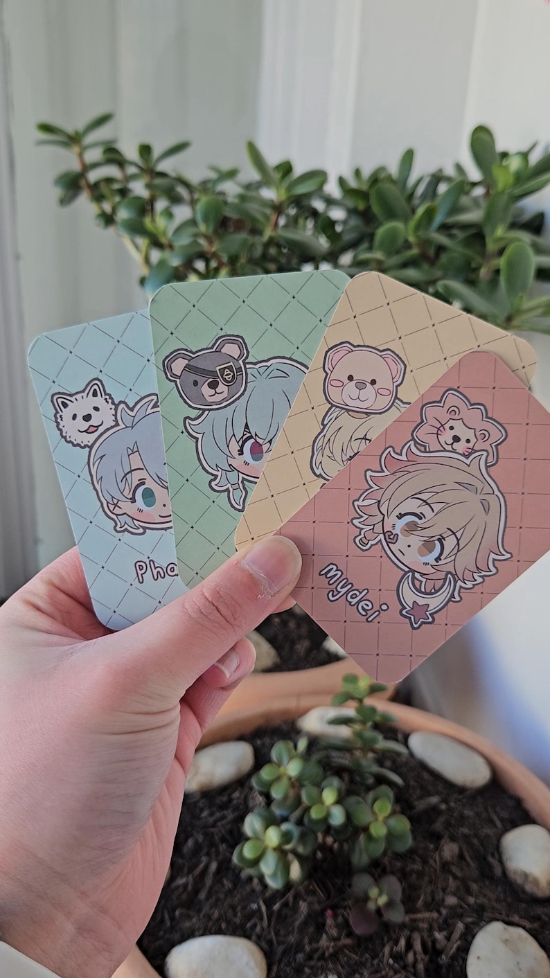 Four back-facing photocards held to the camera. Each card is an illustration of a honkai star rail amphoreus character in a chibi art style with their names labelled at the bottom of the card. From left to right is: Phainon, Anaxa Aglaea and Mydei