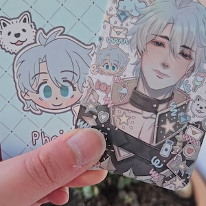 Two of the same photocard are shown. One to display the back-faced card, while the other displays the front face of the card. It is of the character Phainon from honkai star rail. The back-face contains a chibi version of the character while the front is a more detailed illustration of said character decorated with stickers all around them.
