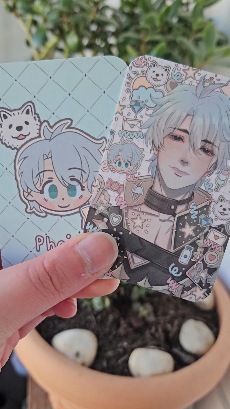 Two of the same photocard are shown. One to display the back-faced card, while the other displays the front face of the card. It is of the character Phainon from honkai star rail. The back-face contains a chibi version of the character while the front is a more detailed illustration of said character decorated with stickers all around them.