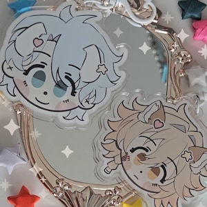 以下が含まれることがあります： 水色とタン色の髪、ハートのアクセント、星のディテールが特徴の漫画のキャラクターデザインが施された2つの透明なアクリルチャーム。チャームは、カラフルな星型の装飾に囲まれた、銀と金のフレームの装飾的な鏡に表示されています。
