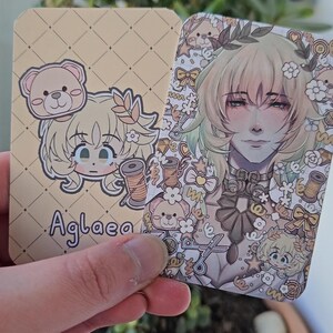 Two of the same photocard are shown. One to display the back-faced card, while the other displays the front face of the card. It is of the character Aglaea from honkai star rail. The back-face contains a chibi version of the character while the front is a more detailed illustration of said character decorated with stickers all around them.