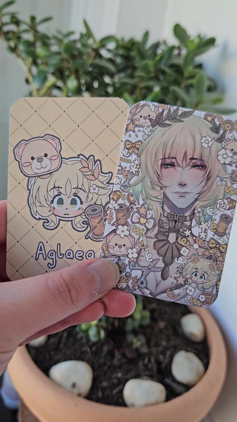Two of the same photocard are shown. One to display the back-faced card, while the other displays the front face of the card. It is of the character Aglaea from honkai star rail. The back-face contains a chibi version of the character while the front is a more detailed illustration of said character decorated with stickers all around them.