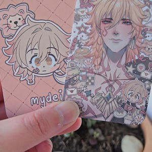 Two of the same photocard are shown. One to display the back-faced card, while the other displays the front face of the card. It is of the character Mydei from honkai star rail. The back-face contains a chibi version of the character while the front is a more detailed illustration of said character decorated with stickers all around them.