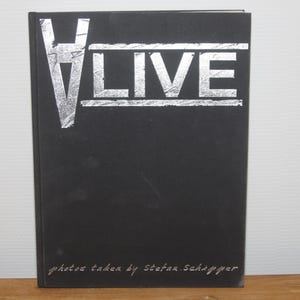Puede incluir: Un libro negro con el texto plateado "V LIVE" en la portada. El texto "fotos tomadas por Stefan Schapper" está impreso en blanco en la parte inferior del libro.