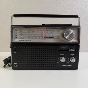 May include: A black and silver Realistic Portavision-5 portable radio with a top handle, a front speaker grille, and a tuning dial. The display panel features AM, FM, TV1, TV2, and VHF bands with a red needle indicator and a headphone jack on the left.