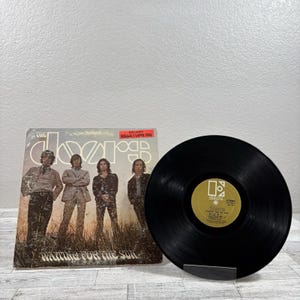 Puede incluir: Portada del álbum "Waiting for the Sun" de The Doors con un disco de vinilo negro. La portada del álbum presenta una foto de la banda y el título del álbum. Una pegatina en la portada dice "Includes Hello, I Love You."