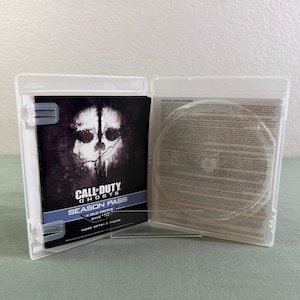 May include: An open clear plastic video game case. The left side holds a Call of Duty Ghosts Season Pass insert featuring a skull graphic and text. The right side contains a software license agreement paper with a circular indentation for a disc.