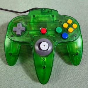 May include: A translucent green video game controller featuring a gray directional pad, a central gray analog stick, a red Start button, a green B button, a blue A button, and four yellow C-buttons. The Nintendo logo is embossed at the top.