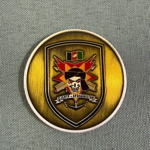 Puede incluir: Una moneda de desafío circular en tono latón con un emblema central en forma de escudo. El diseño incluye una calavera con un pañuelo oscuro, una estrella roja y amarilla, flechas cruzadas, un ancla y el texto CJSDTF - AFGHANISTAN.