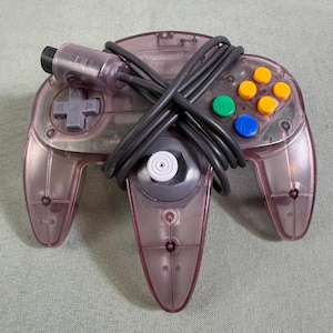 May include: A translucent purple video game controller featuring a gray directional pad, a central gray analog stick, and four colored action buttons in green, yellow, and blue. A black connection cable is wrapped around the controller body.