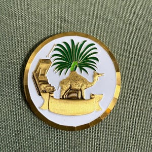 Op de afbeelding: Een ronde metalen munt in goud en wit met een gouden kameel onder een groene palmboom. Links van de kameel staat een gouden tank en onder de kameel een leeg gouden lint. De munt heeft een gepolijste gouden rand.