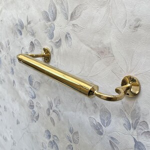 May include: A polished brass towel bar mounted on a floral wallpapered wall. The bar is cylindrical with curved support arms and circular mounting plates. The brass finish reflects light, creating a bright accent.