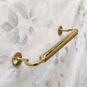 May include: A polished brass handrail, mounted on a wall with a floral pattern. The handrail has a cylindrical bar with curved supports and circular mounting plates. The brass finish reflects light, creating a bright, elegant look.