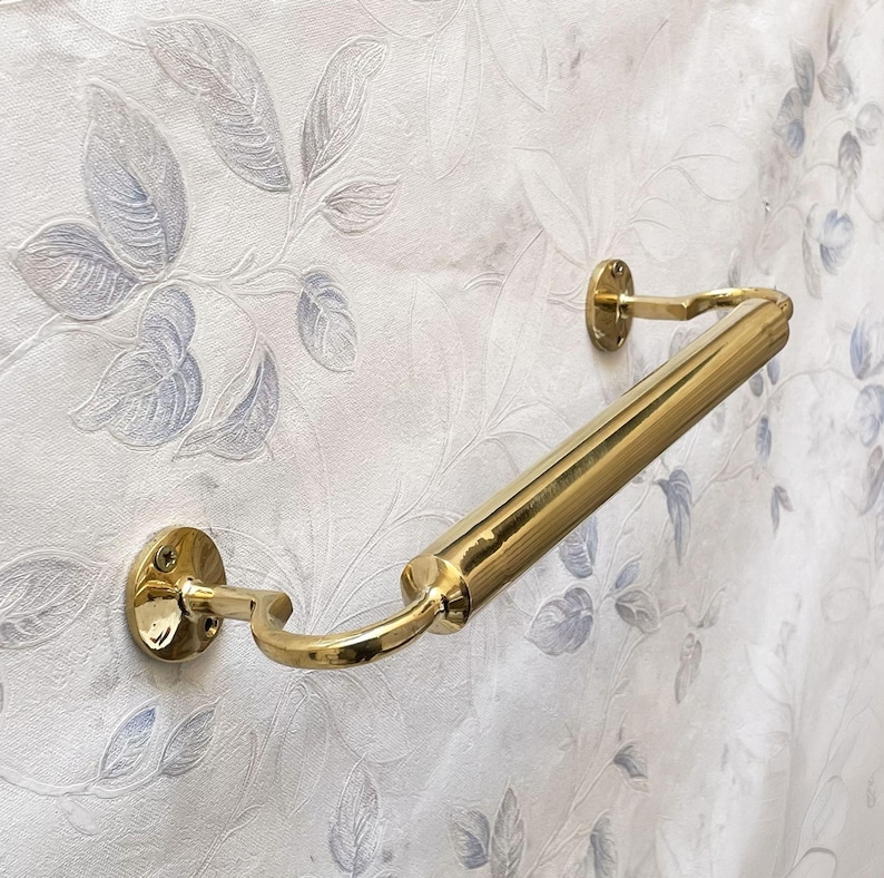 May include: A polished brass handrail, mounted on a wall with a floral pattern. The handrail has a cylindrical bar with curved supports and circular mounting plates. The brass finish reflects light, creating a bright, elegant look.