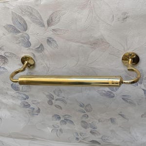 May include: A gold-colored towel bar with a cylindrical center section and curved end supports. The bar is mounted on circular plates, and the design is simple and elegant. The background features a floral pattern.