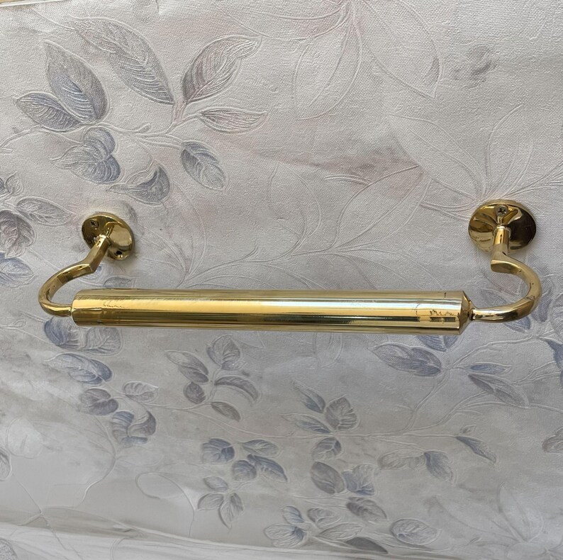 May include: A gold-colored towel bar with a cylindrical center section and curved end supports. The bar is mounted on circular plates, and the design is simple and elegant. The background features a floral pattern.