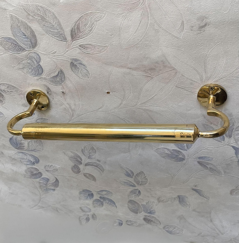 May include: A gold-colored towel bar with curved ends and circular mounting plates. The bar is cylindrical and appears to be made of metal. The background features a floral pattern in shades of gray and white.