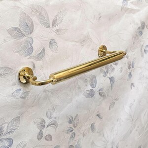 May include: A polished brass towel bar with a cylindrical center section and curved end supports. The bar is mounted on a wall with a floral pattern in shades of gray and white. The bar is approximately 18 inches long.