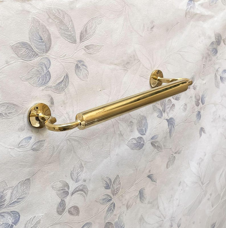 May include: A polished brass towel bar with a cylindrical center section and curved end supports. The bar is mounted on a wall with a floral pattern in shades of gray and white. The bar is approximately 18 inches long.