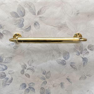 May include: A gold-colored towel bar with a cylindrical design and rounded ends. The bar is mounted on a wall with a floral pattern in shades of gray and white. The bar is approximately 18 inches long.