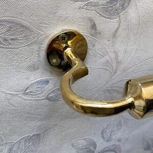 May include: A close-up of a brass-colored hook attached to a wall. The hook has a circular base with a screw and a curved arm. The wall has a white and gray floral pattern.