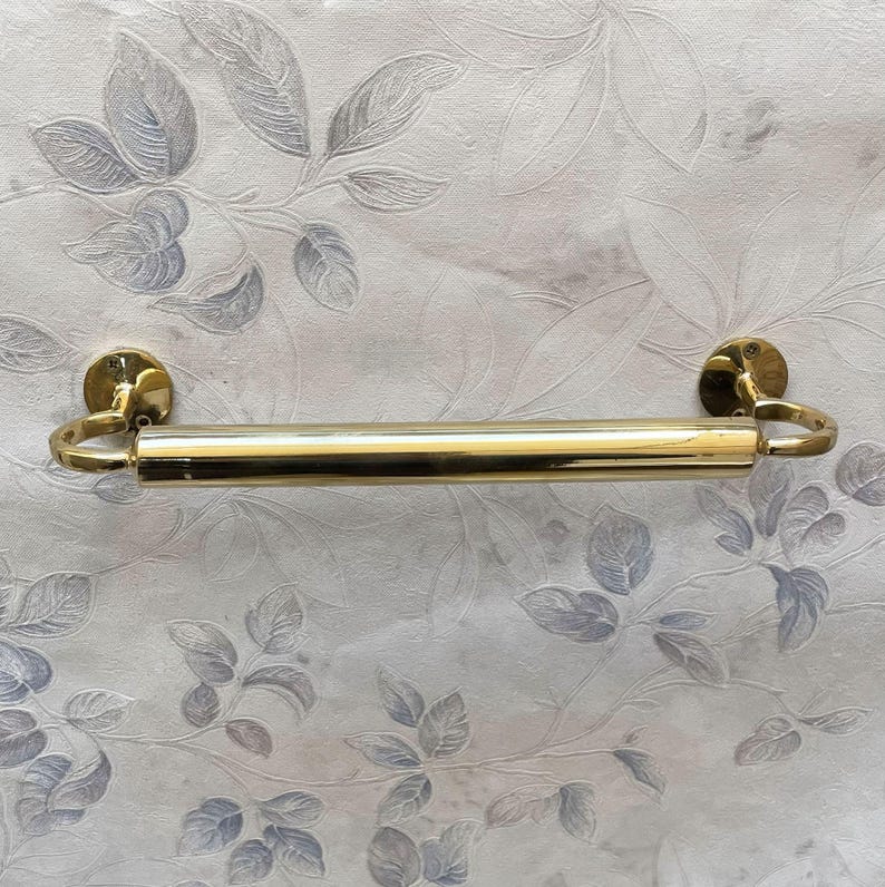 May include: A gold-colored towel bar with a cylindrical design. The bar is mounted on two circular wall plates with curved, ring-shaped accents. The background features a floral pattern in shades of gray and white.
