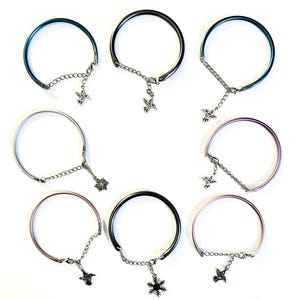 May include: Collection of seven metal bracelets in various colors, including blue, silver, and pink. Each bracelet features a chain and a decorative charm, such as a hummingbird or snowflake. The bracelets are displayed on a white background.