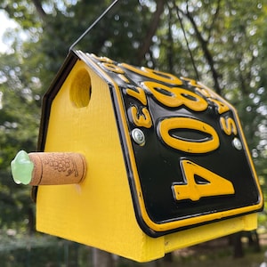 Può includere: Una casetta per uccelli gialla e nera realizzata con una targa riutilizzata con il numero "408" su di essa. La casetta per uccelli ha un piccolo mangiatoia per uccelli verde attaccato al lato.