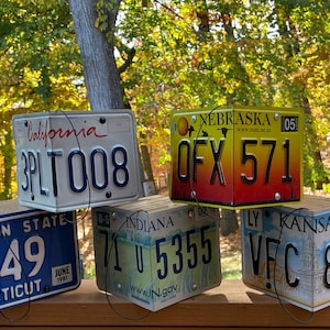 Puede incluir: Cinco placas de matrícula de diferentes estados están dispuestas en forma de cuadrado. Las placas son de California, Nebraska, Connecticut, Indiana y Kansas. Las placas son todas de diferentes colores y tienen diferentes diseños.