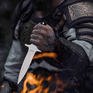 May include: A silver dagger with a black handle is held. The dagger has a double-edged blade and a crossguard. The person is wearing armour with intricate designs. Flames are visible in the foreground. The hand holding the dagger has tattoos.