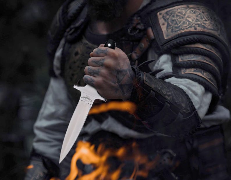 May include: A silver dagger with a black handle is held. The dagger has a double-edged blade and a crossguard. The person is wearing armour with intricate designs. Flames are visible in the foreground. The hand holding the dagger has tattoos.