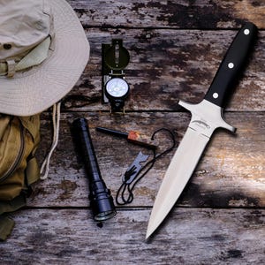 May include: A collection of outdoor survival gear including a knife with a black handle and silver blade, a compass, a torch, a fire starter, a beige hat, and a backpack. The items are arranged on a weathered wooden surface.